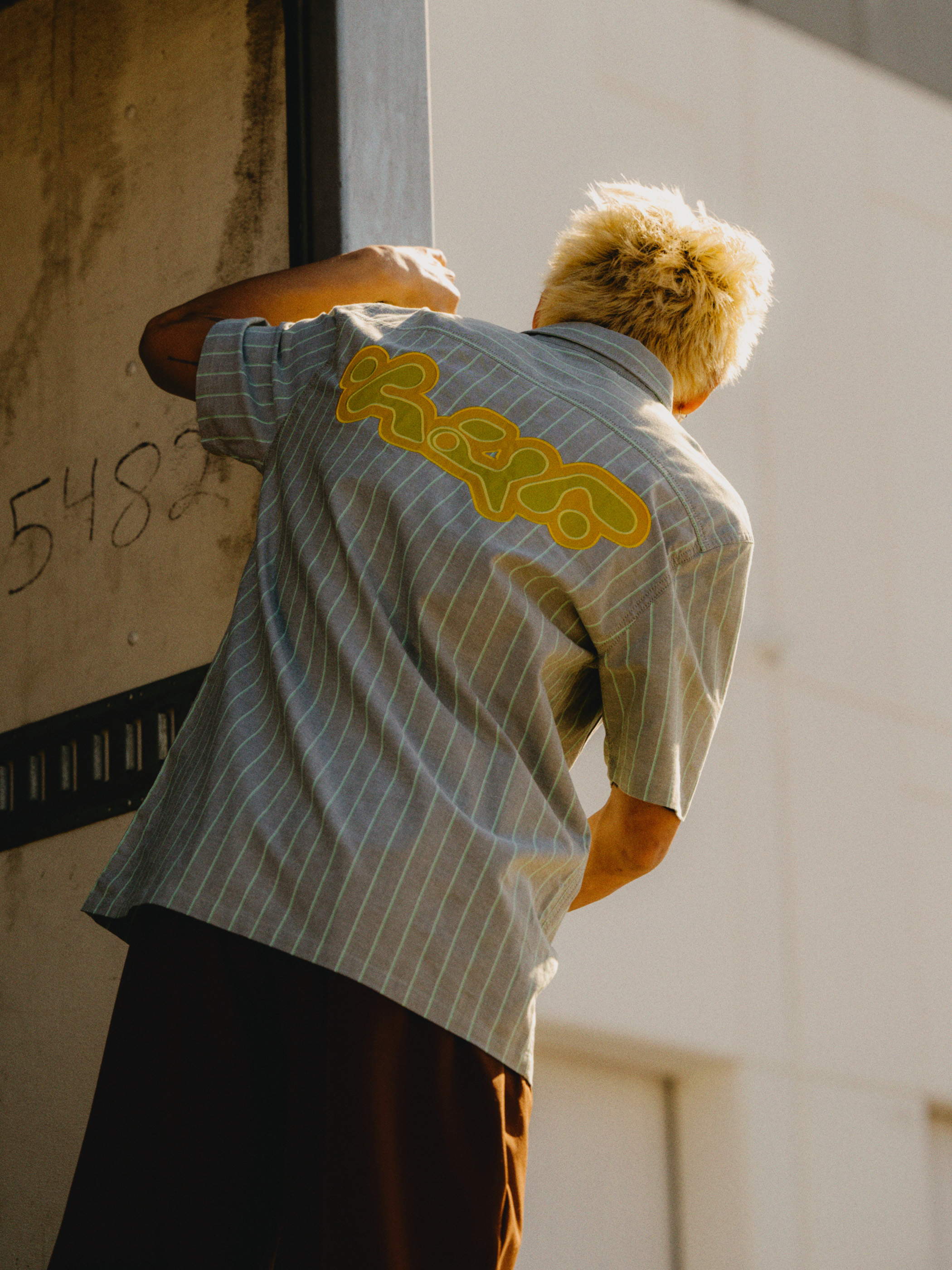 Person wearing striped shirt with yellow logo on back, brown pants, fair skin, blond hair, stands near gray wall in daylight.