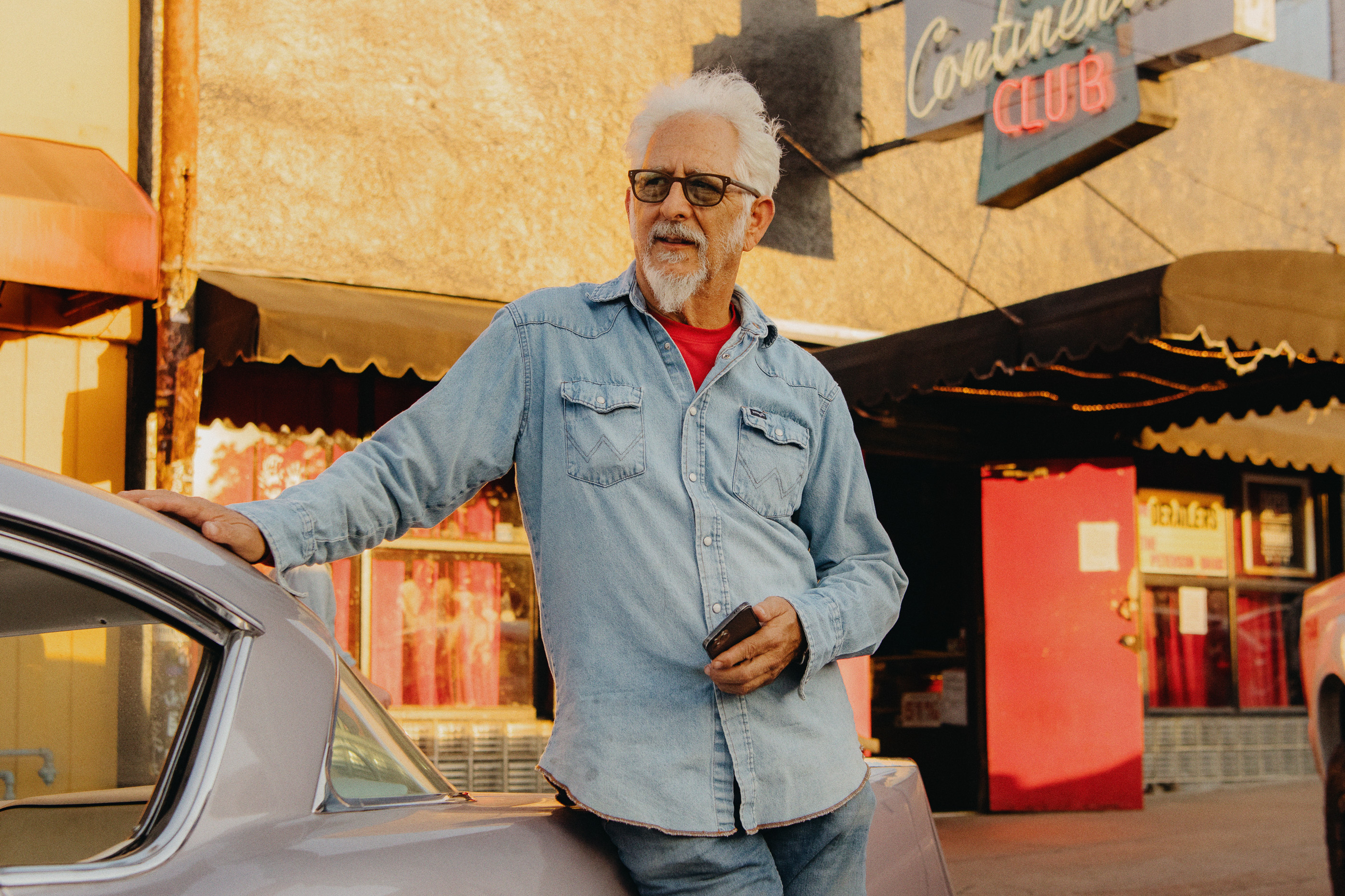 Elderly man with white beard and glasses wearing a blue denim shirt leaning on a car outside a club in Los Angeles
