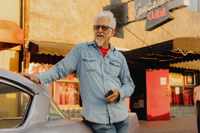 Elderly man with white beard and glasses wearing a blue denim shirt leaning on a car outside a club in Los Angeles