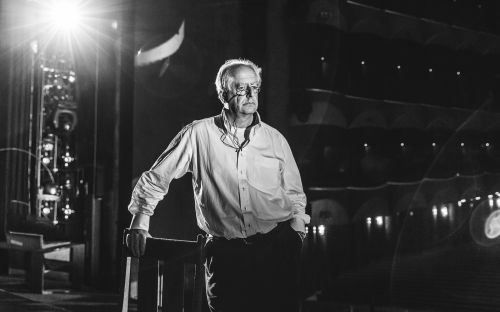 Monochrome portrait of an elderly man with glasses wearing a button-down shirt standing and leaning on a chair in a dark venue.