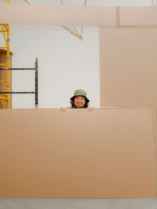 Smiling man with a bucket hat peeks over a cardboard wall, positioned in an indoor setting with a scaffold visible in the background.