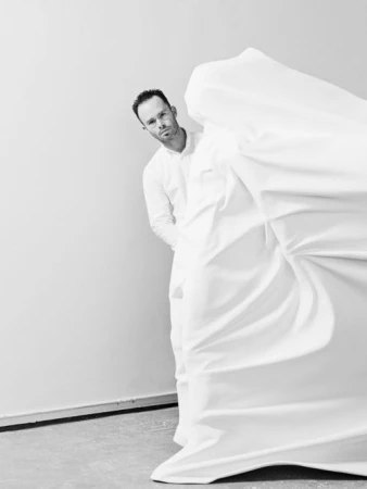 Man wearing a white shirt standing beside a large draped white fabric sculpture in a studio