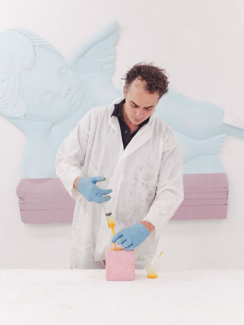 A man wearing a white coat and blue gloves injects yellow liquid into a pink block in a white interior with a light-blue sculptural background.