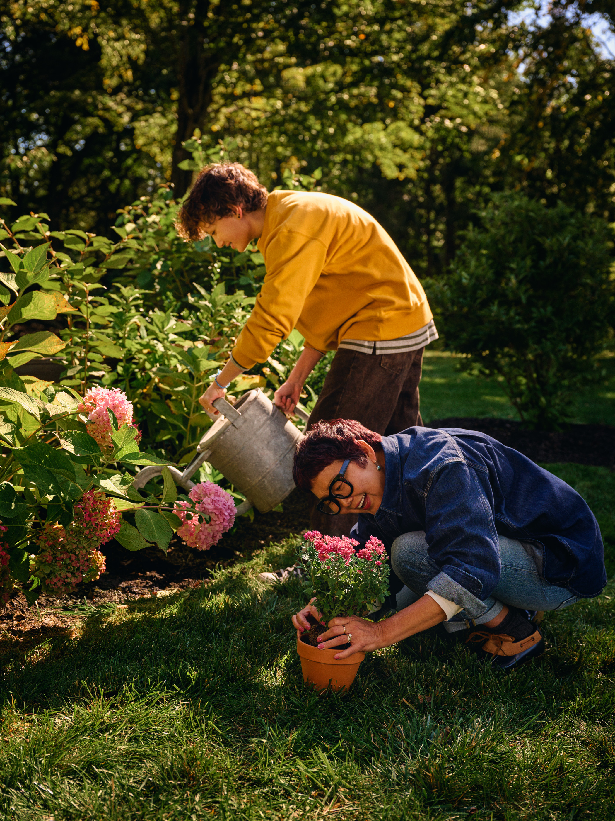 Two people gardening in a lush outdoor garden setting with a variety of plants, flowers, and trees on a sunny day.