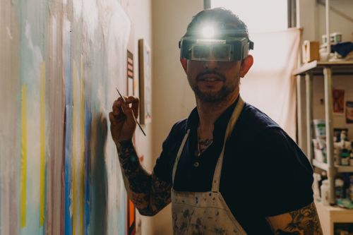 Adult male artist painting on a large canvas in an art studio, wearing a headlamp and apron, showcasing his tattooed arms.