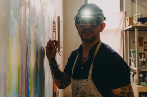 Adult male artist painting on a large canvas in an art studio, wearing a headlamp and apron, showcasing his tattooed arms.
