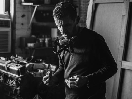 A man in a workshop wearing a mask and holding small metal parts, with an engine and tools visible in the background in black and white.