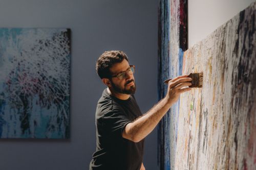 An artist with glasses is painting on a large canvas in an indoor art studio setting, using a wide brush and wearing a black t-shirt.