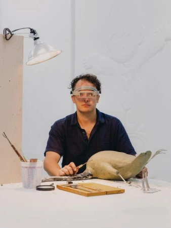 Adult male artist wearing magnifying glasses meticulously carves a bird sculpture in a bright studio, using hand tools.