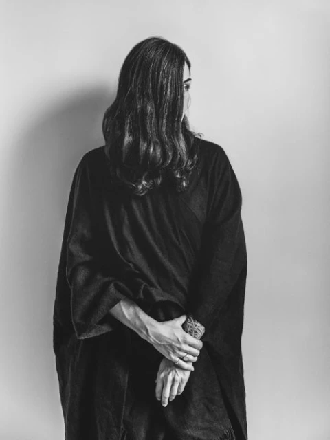 Black and white portrait of a woman with her back to the camera, looking over her shoulder with her hands clasped at her waist.