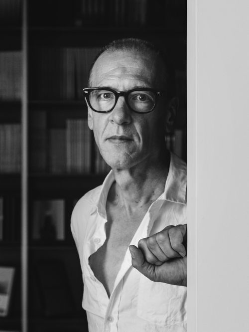 Black and white portrait of an adult man wearing glasses and an unbuttoned shirt, standing in an indoor environment with a bookshelf visible.