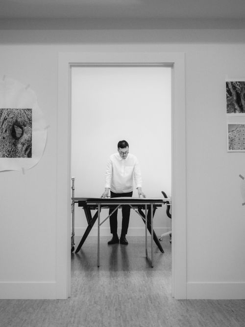 Man wearing a white shirt standing at a dark drafting table inside a room framed by a white doorway