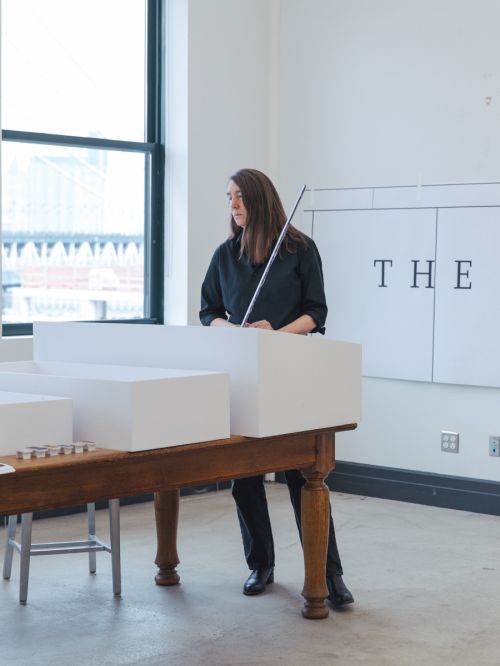 Adult female standing indoors at a wooden table with white foam blocks, holding a metal rod, near a bright window and signage.