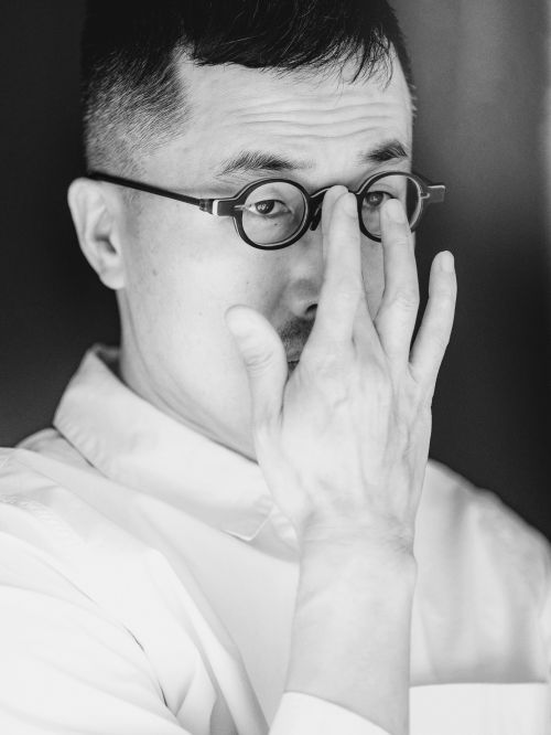Monochrome portrait of a man with glasses holding his hand up to his face in an indoor environment with a dark background.