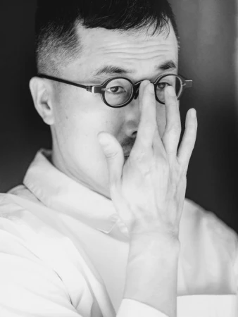 Monochrome portrait of a man with glasses holding his hand up to his face in an indoor environment with a dark background.