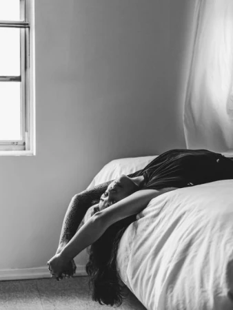 Black and white portrait of a woman lying backward on a bed with arms extended near a window by photographer Dustin Aksland