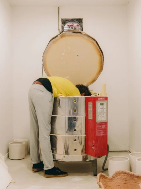 Adult male bends over a large silver and red kiln in a well-lit white room, surrounded by ceramic pots and pottery.