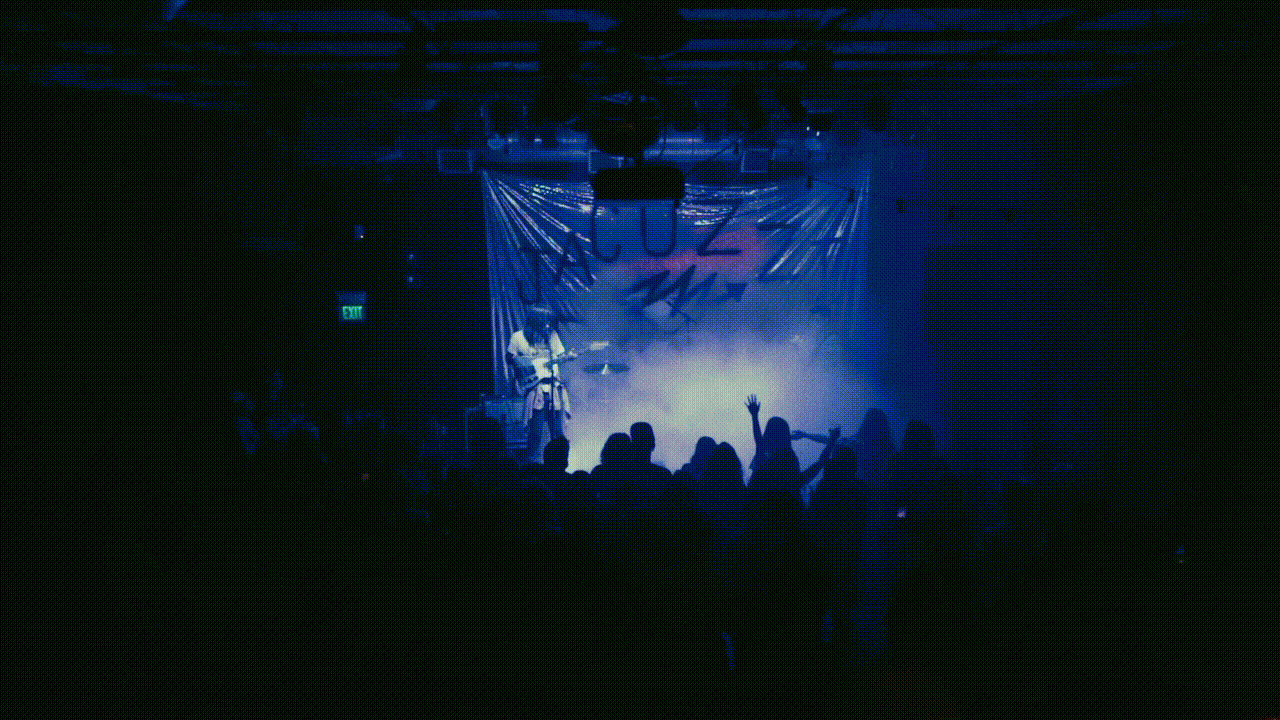 Live music performance with band on stage in blue lighting, silhouetted crowd visible in foreground, banner in background.