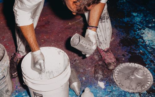 Overhead shot: person wearing gloves pours paint-like liquid into a white bucket, surrounded by paint containers and multicolored floor.