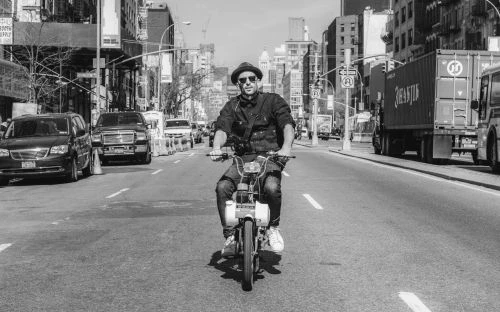 A monochrome image features a man riding a motorcycle on an urban street, flanked by tall buildings and various vehicles.