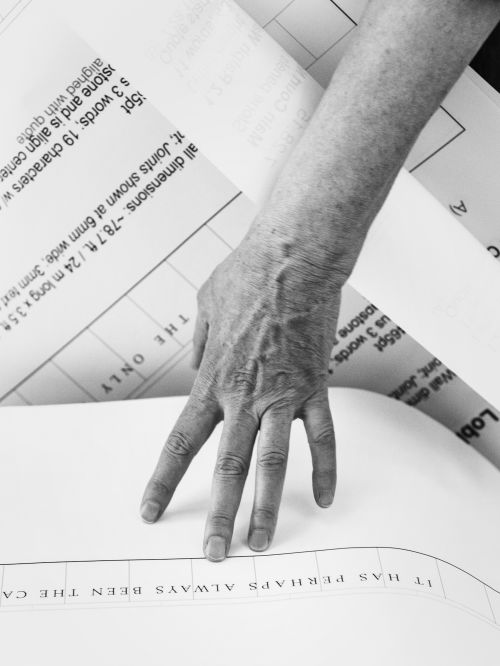 Close-up of a hand touching blueprint on a white surface. Architectural plans are in background. Black and white photography.