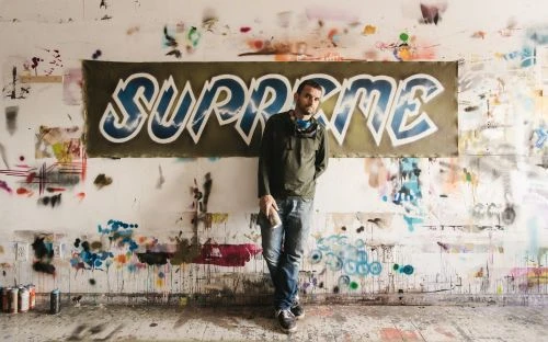 Man holding a spray can leaning against a paint-splattered wall featuring a large banner reading SUPREME, photographed by Dustin Aksland.