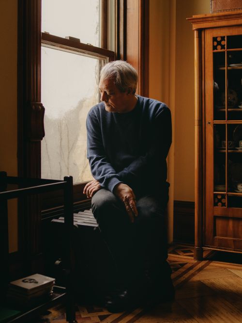 Elderly man seated indoors by window, wearing a navy sweater and dark trousers, with a vintage cabinet and parquet floor visible.