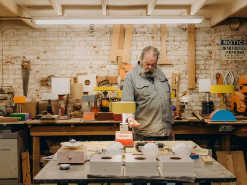 Man crafting colorful wooden lamps in a workshop with tools, painted wood, and a brick wall backdrop in a brightly lit workspace.