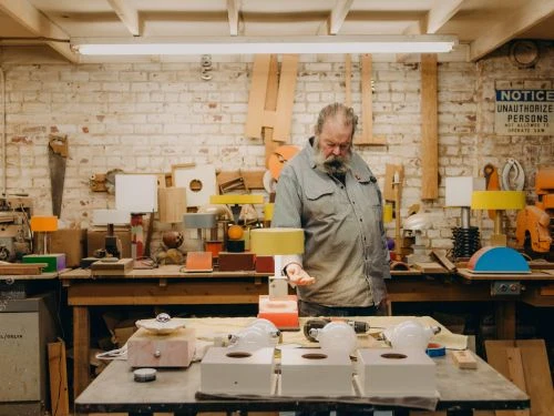 Man crafting colorful wooden lamps in a workshop with tools, painted wood, and a brick wall backdrop in a brightly lit workspace.