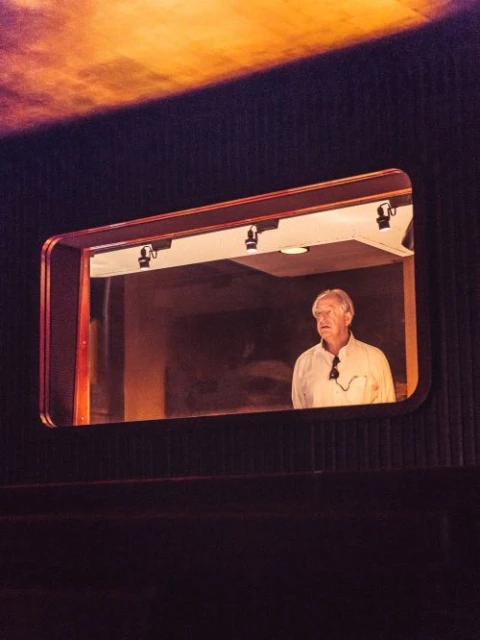 A man with white hair stands in a room, framed by a rectangular opening with lights above, viewed from a theater-like setting.