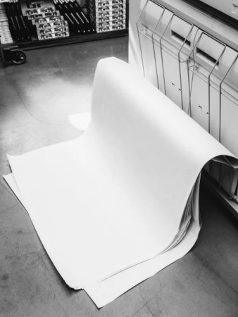 Stack of oversized white paper sheets on the floor near a printing press in black and white, indoor industrial setting, overhead angle.
