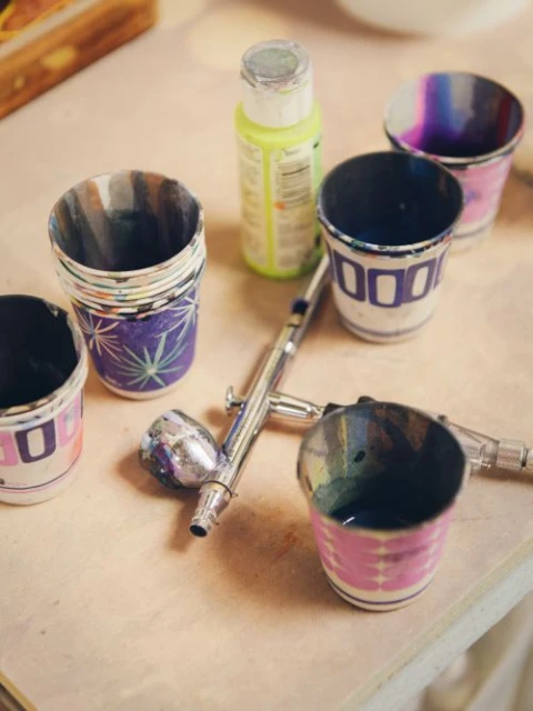 Close-up of an airbrush gun, multiple paint cups with colorful paint residue, and a bottle of green paint on a light-colored surface.