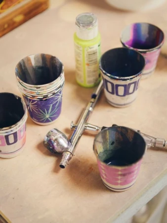 Close-up of an airbrush gun, multiple paint cups with colorful paint residue, and a bottle of green paint on a light-colored surface.