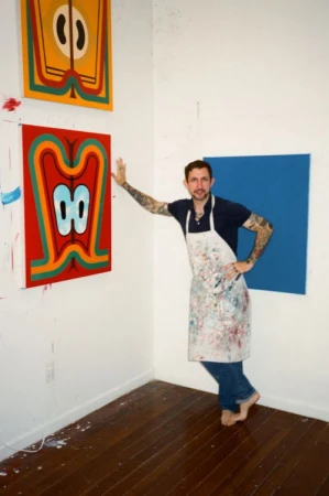 Adult male artist in a navy shirt, jeans, and paint-covered apron poses barefoot in an art studio with two abstract paintings.