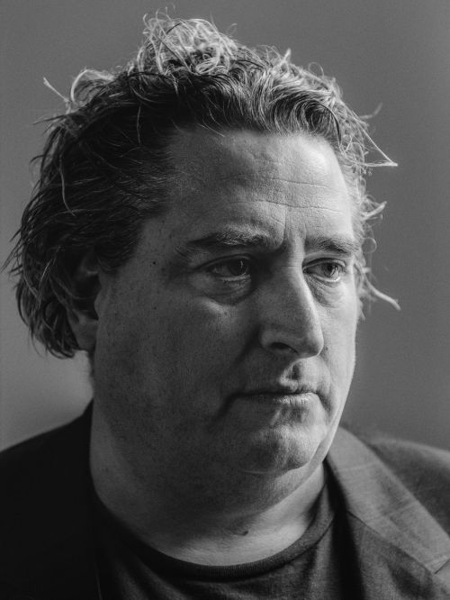 Black and white close-up portrait of an adult male with light colored, slightly messy hair wearing a dark coat and t-shirt.