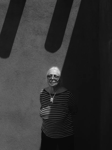 Black and white portrait of a mature woman with eyeglasses wearing a striped shirt standing against a wall with light and shadow.