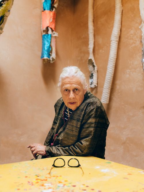 Elderly woman with white hair and glasses sits indoors with a patterned table. She looks at the camera with a neutral expression.