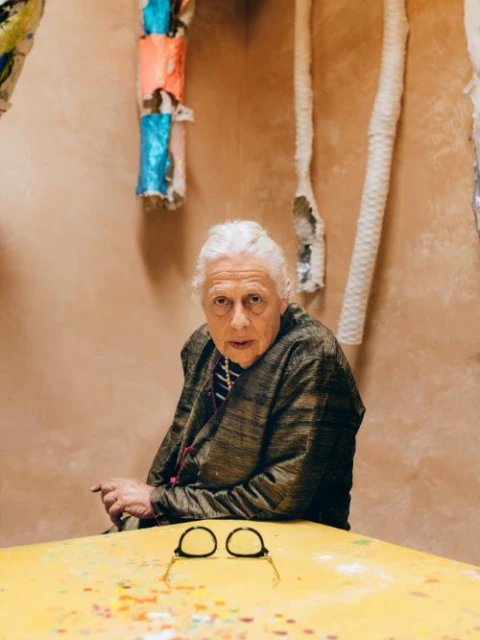 Elderly woman with white hair and glasses sits indoors with a patterned table. She looks at the camera with a neutral expression.