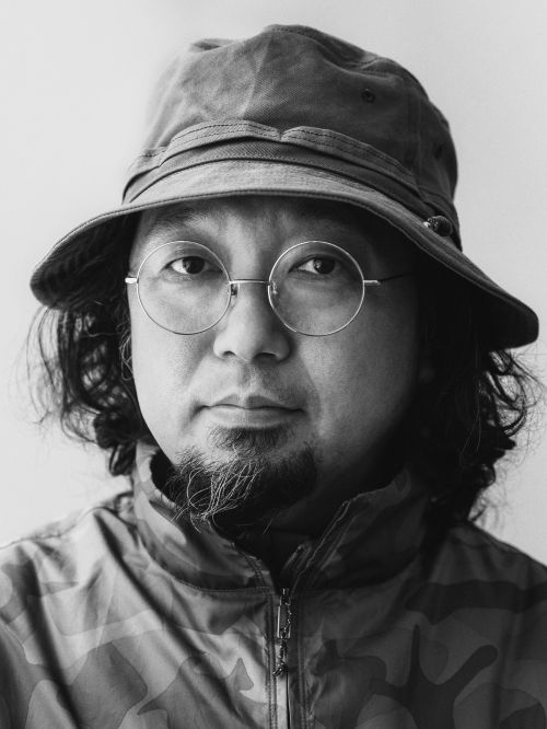 Black and white portrait of a man wearing round glasses, a patterned bucket hat, and a zippered jacket against a plain wall.