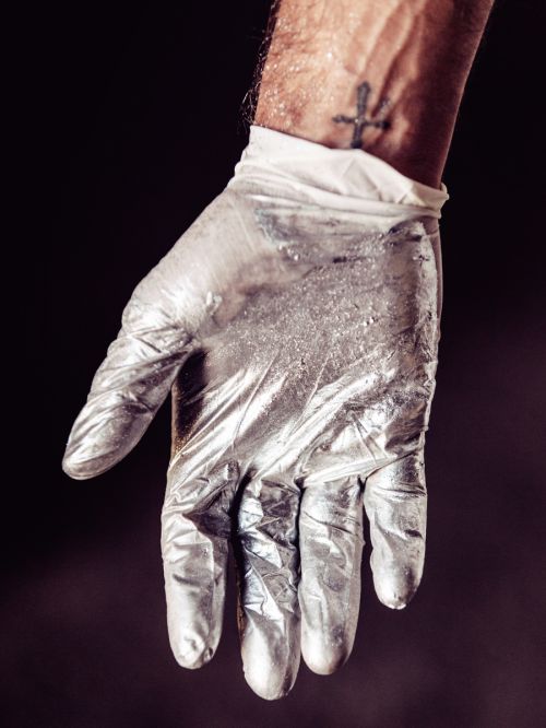 Close-up shot of a gloved hand with silver paint and an arm with a tattoo against a dark background, creating a mysterious artistic image.