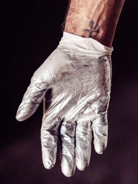Close-up shot of a gloved hand with silver paint and an arm with a tattoo against a dark background, creating a mysterious artistic image.