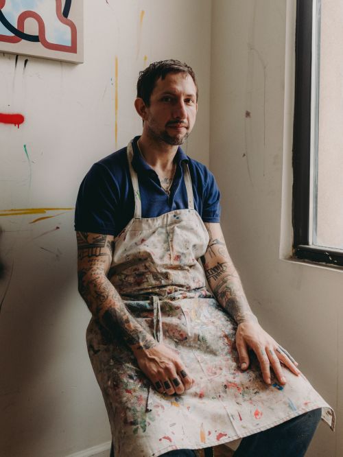 Indoor portrait of a fair-skinned adult male artist sitting in front of a painting with a paint-splattered apron in a bright room.