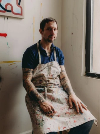 Indoor portrait of a fair-skinned adult male artist sitting in front of a painting with a paint-splattered apron in a bright room.