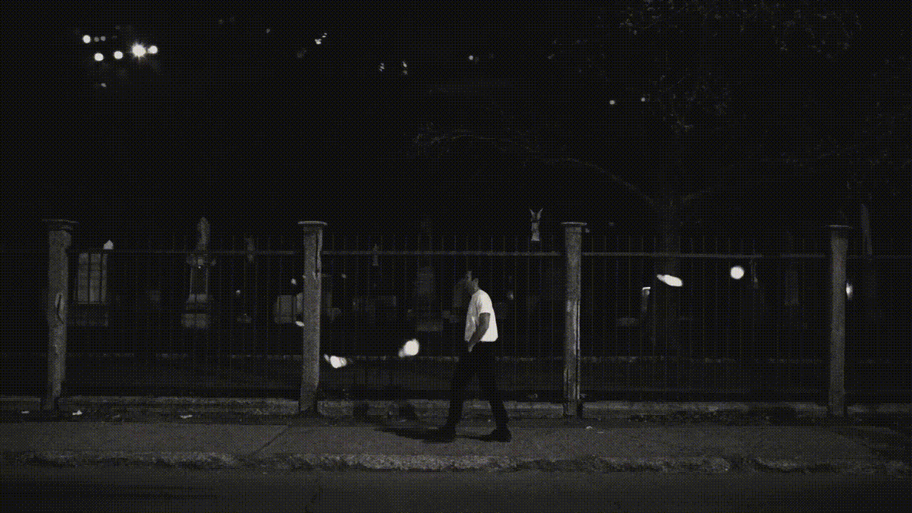 A man walks past a fence in a black-and-white nighttime scene, illuminated by street lights and moonlight, wearing a light shirt.