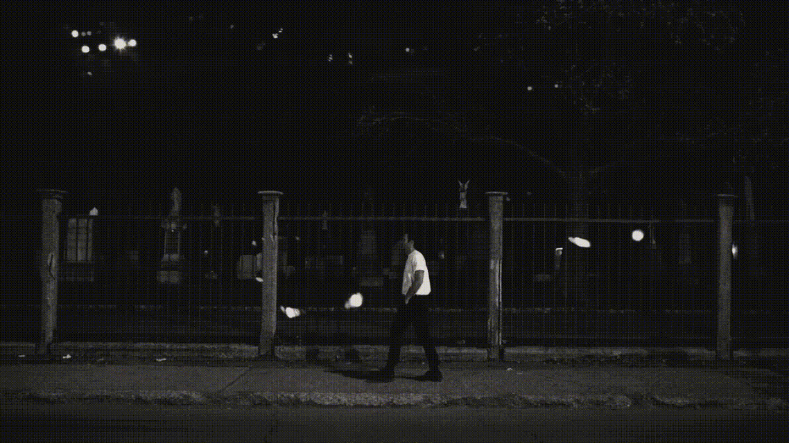 A man walks past a fence in a black-and-white nighttime scene, illuminated by street lights and moonlight, wearing a light shirt.
