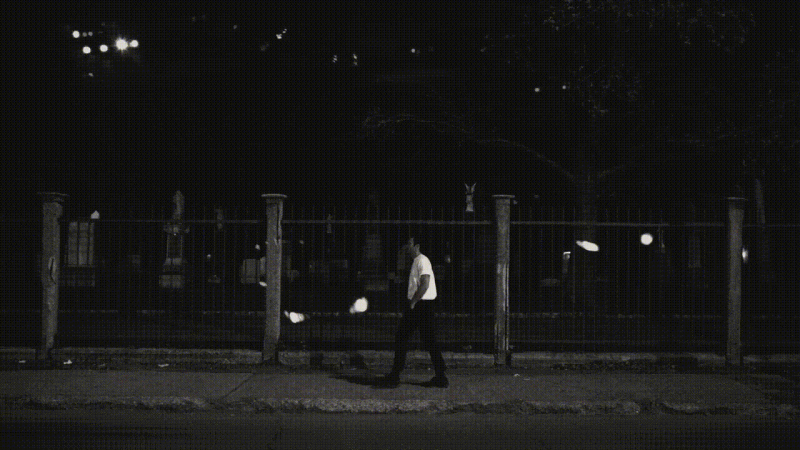 A man walks past a fence in a black-and-white nighttime scene, illuminated by street lights and moonlight, wearing a light shirt.