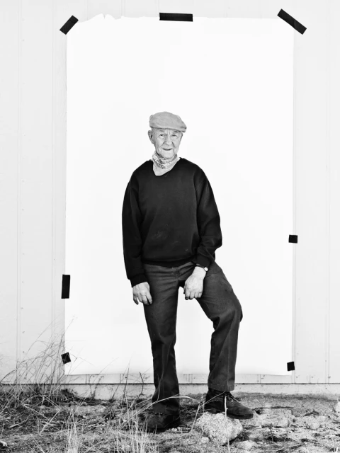 Monochrome portrait of an elderly man in cap, sweater, jeans posing outdoors in front of white backdrop, standing on stone.
