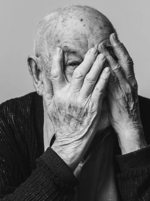 Elderly man covers his face with wrinkled hands in a black-and-white portrait. The image highlights aging, skin texture, and human emotion.