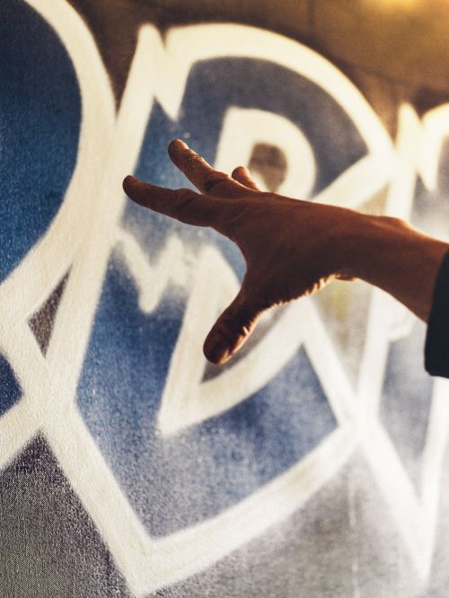 Close-up of a hand gesturing toward a white and blue abstract graffiti design painted on a textured gray wall surface indoors.