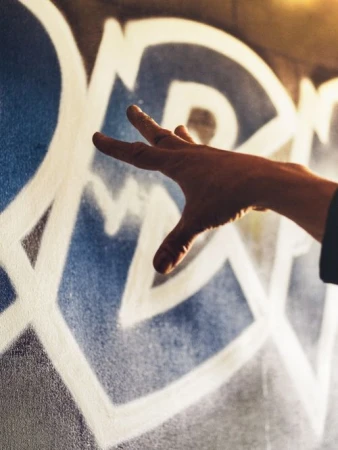 Close-up of a hand gesturing toward a white and blue abstract graffiti design painted on a textured gray wall surface indoors.